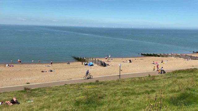  Panning shot of whitstable Beach harbour in summer on hot day with sunshine people walking on beach amd cyclist riding on pathment sunbathing