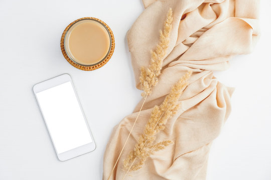 Modern minimal home office desk with beige blanket, cup of coffee, dry flowers and smartphone with blank screen mockup. Hygge or boho style women's workspace. Top view, flat lay.