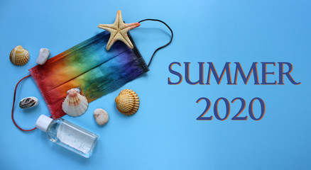 Summer vacation flat lay with sea shells, pebbles and starfish next to protective mask decorated in rainbow colors and antiseptic on blue background. Travel and coronavirus covid-19. Summer 2020 sign