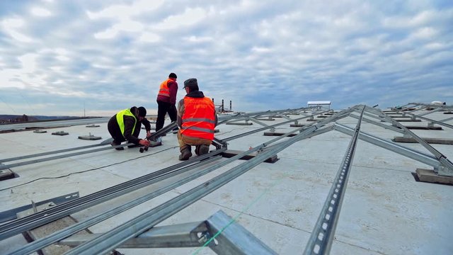 Solar panel technicians on flat roof of a big building. Metal base before the construction of photovoltaic panels. Workers installing new solar farm.