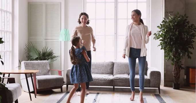 Full length overjoyed multigenerational family celebrating special occasion, dancing barefoot in living room. Active middle aged woman having fun with grown daughter and small granddaughter indoors.
