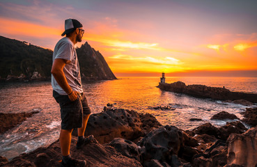 A young man in a cap looking at the orange sunset of the Pasajes San Juan lighthouse, near San...