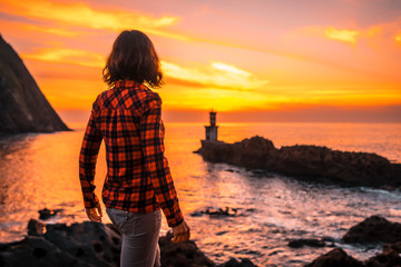 A young woman watching the orange sunset from the Pasajes San Juan lighthouse, near San Sebastian,...