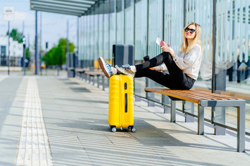 Tired blond woman traveler sitting on bench at bus or tram stop, holding puting legs on her...