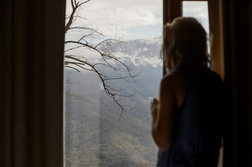Blurred silhouette of a girl who watches the mountain landscape outside the window. Snow-capped peaks and green forests of the Caucasus Mountains.