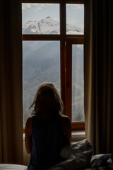 A girl sits on a bed in her room in front of the window and admires the morning mountain landscape. She delight in view of snow-capped peaks and green forests of the Caucasus Mountains.