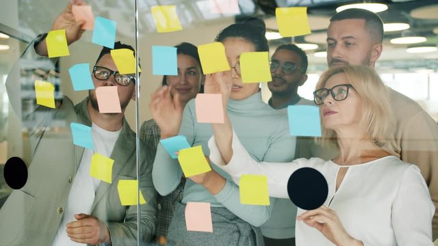 Business people solving business problems in office using sticky notes on glass board sticking multicolored paper with information working together.