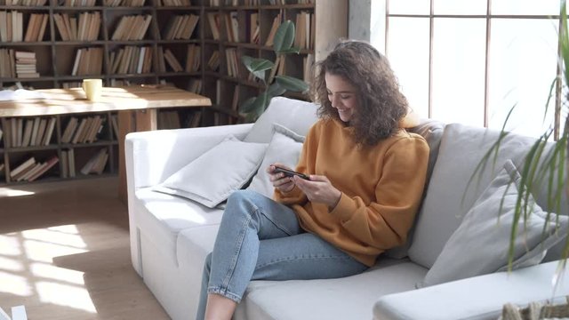 Smiling young hispanic latina gen z teen girl sit on sofa at home holding phone watching funny social media videos looking at mobile screen, laughing, relaxing in cozy living room using cellphone.