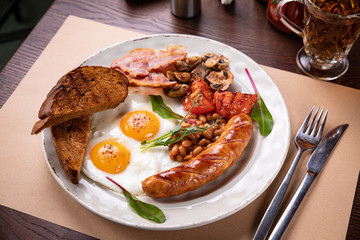 Full English breakfast - fried egg, baked beans, bacon, sausages on a dark rusty background, toasts.