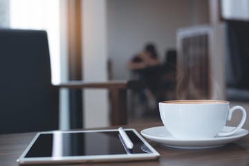 Cup of coffee and digital tablet on wooden table in loft style coffee shop coworking space