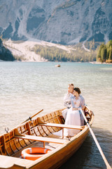 Fototapeta na wymiar A wedding couple is sitting in a wooden boat on the Lago di Braies in Italy. Newlyweds in Europe, on Braies lake, in the Dolomites. The groom hugs the bride.