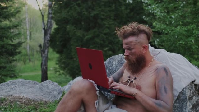 Ancient Prehistoric Caveman Using Laptop Computer in Forest. Brutal Serious Bearded Man Typing text or Programming. Distance or Remote Work, Freelancer or Business Startup.