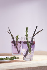 Violet cocktail with ice cubes is contained in a glass with reusable black metal straws, a sprig of rosemary and raspberries. The glass is located near a white plate with herbs on the wooden table.