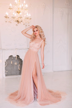 Young beautiful sexy woman fashion model posing. Hairstyle girl graduate wavy blonde hair hoop band pearl accessories. Luxury beige peach pink nude dress, open bare leg. Backdrop white room classic