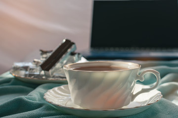 A cup of tea on the bed and light blue blanket with sunrise, Morning teatime with chocolate, notebook computer,smartphone, Normal practice by working at home during covid-19 outbreak around the world.