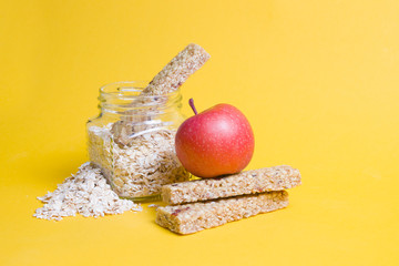 glass jar with oatmeal, an apple and several protein bars for a snack on a yellow background, sprinkled oatmeal, copy space, healthy proper nutrition concept