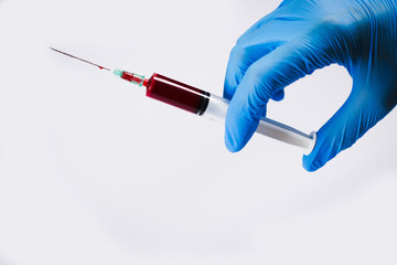 Hand with blue nitrile glove is holding syringe with red liquid, in front of white background....