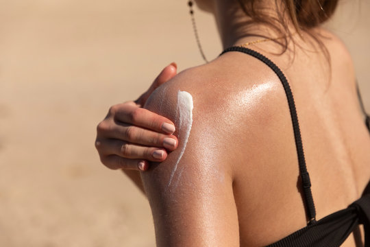 A young woman applying sun cream or sunscreen on her tanned shoulder to protect her skin from the sun. Shot on a sunny day with blurry sand in the background.
