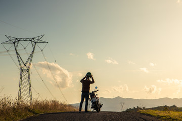 Man standing next his motorbike taking off helmet along a country road on hills landscape at sunset. Tuscany, Italy.