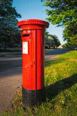 Fototapeta na wymiar Iconic red british Royal Mail post box in the evening sunshine on a quite road with a green tree lined verge