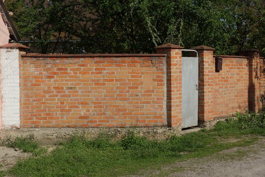 red brick wall of the fence and gray iron door on the street in green grass and vegetation