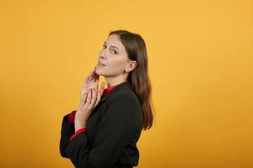 Young Attractive Woman Brunette In Black Stylish Suit, Red Shirt On Yellow Background, Gentle Female Holds Hands To Her Face. The Concept Of Kind, Nice People