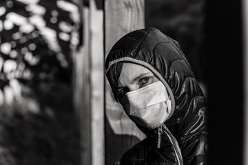 Portrait of a sad young woman in a medical mask on her face and a hood in nature alone. Dark picture of loneliness during the virus.