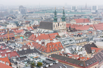 Naklejka na ściany i meble Cityscape with red tiles rooftops and tower of Jesuit Church of the old town of Vienna in a heavy snowy day. View at the tower of St. Stephen's Cathedral in Vienna, Austria.