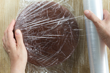 Top view of woman hand wrapping chocolate sponge cake in plastic for storing on the wooden background