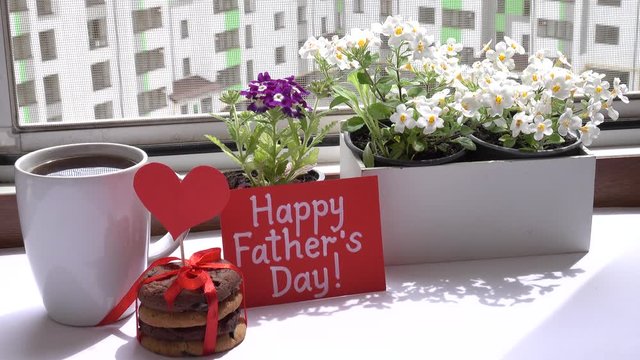 A greeting card in honor of Father's Day is standing near primrose flowers, a cup of hot tea and a holiday cookies on a white table near open window in sunny day