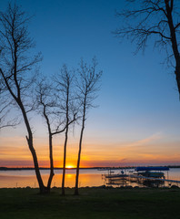 Sunset over the lake with trees, dock, lifts, and a boat in the foreground in silhouette.