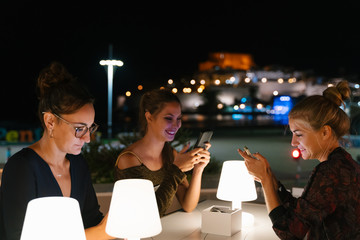 Three young women sitting on a terrace at night while using their mobile