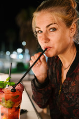 Blonde woman looking at the camera and drinking a red cocktail with a straw on a terrace at night