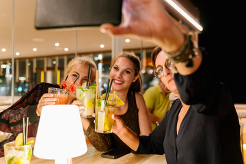 Frontal photo of women making a selfie while sitting on a terrace drinking cocktails at night