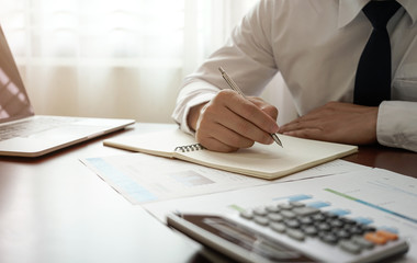 Close up of businessman in shirt and tie working in his office writing in notebook on table with calculator, laptop, document.