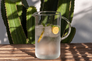 A carafe with ice water, slices of lemon and mint leaves is on a wooden table against a background...