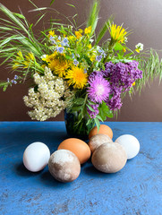 Multi-colored Easter eggs and bright wild flowers in a decorative vase, against a background of an abstract surface. Free space.