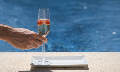 Man takes a glass with champagne or prosecco with raspberry on swimming pool or sea blurred background. Happy white man put glass on poolside in the sunny day. Luxury drink.