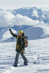 Girl  bright helmet and backpack on a snowboard admires snow-capped mountain peaks and clouds which hanging low overhead. She is sending the greetings to camera with happy smile and with hands up. 