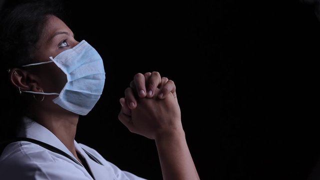 Profile view of young woman doctor in medical mask praying to god on black background looking up - concept of hope and fight to end coronavirus or covid-19 crisis.