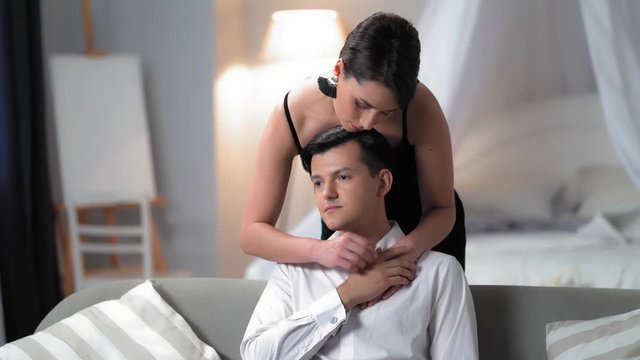 Charming Young Woman In A Black Dress Is Standing Behind The Couch and Hugging Gently A Man In A White Shirt Sitting On The Sofa. The Couple Is In The Stylish Bedroom.