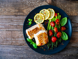 Fish dish - fried cod fillet with asparagus on wooden table  - obrazy, fototapety, plakaty