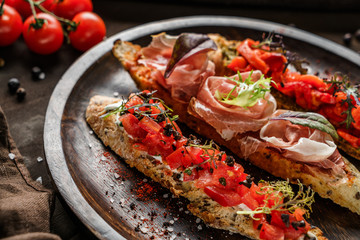 Variety of bruschetta with prosciutto, tomatoes, parmesan cheese, sauce pesto, grilled pepper on ciabatta bread on wooden plate over dark background. Sandwiches set of wine, top view