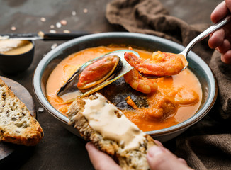 Tomato gazpacho soup with mussels, squid, shrimp in bowl over dark wooden background. Hands holding a spoon with prawn and soup. Seafood, healthy food, close up