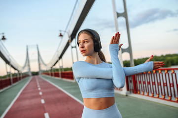 A beautiful athlete girl warming-up before outdoor workout. Girl in sports suit is stretching her arms and listening music in a headphones on a city bridge