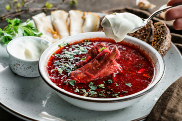 Ukrainian and Russian traditional beetroot soup - borscht in bowl with rib eye meat, rye bread, slices of bacon on wooden background. Hand holds a spoon with sour cream. Healthy food, close up