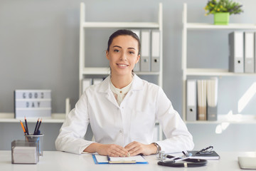 Fototapeta premium Positive doctor woman looking at the camera smiling while sitting at a table in a clinic.