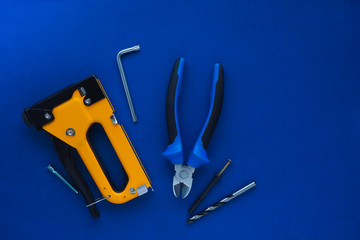 black and yellow construction stapler, pliers, drill, furniture key and screws on a blue background. flat layout, top view. Space for text.