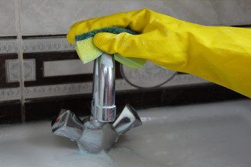A faucet in the bathroom is being cleaned by a female hand in a yellow, rubber glove. To clean the chrome tap use a powder and a sponge. The concept of cleaning the sink and cleaning the bathroom.