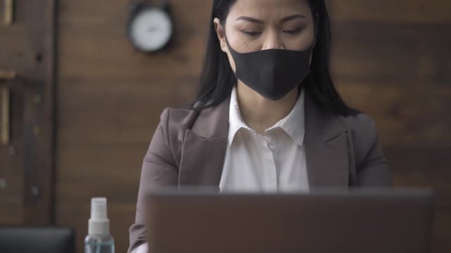 Businesswoman in pollution mask in front of a laptop working in a coronavirus quarantine days from office with wooden background. High quality footage